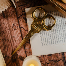 Load image into Gallery viewer, Top view of golden skull scissors set against a wooden table and page of a book