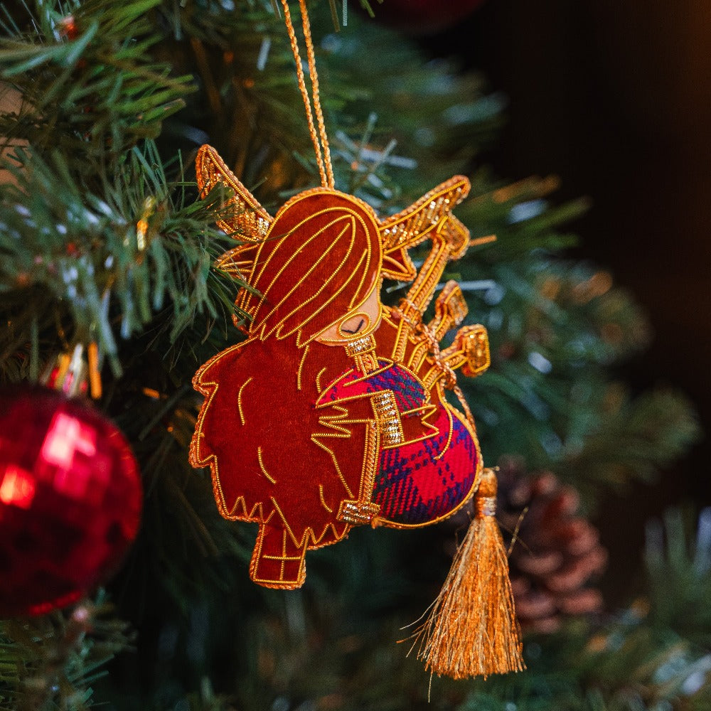 Hand-stitched bagpiping cow decoration hanging on a Christmas tree with a large, gold tassel