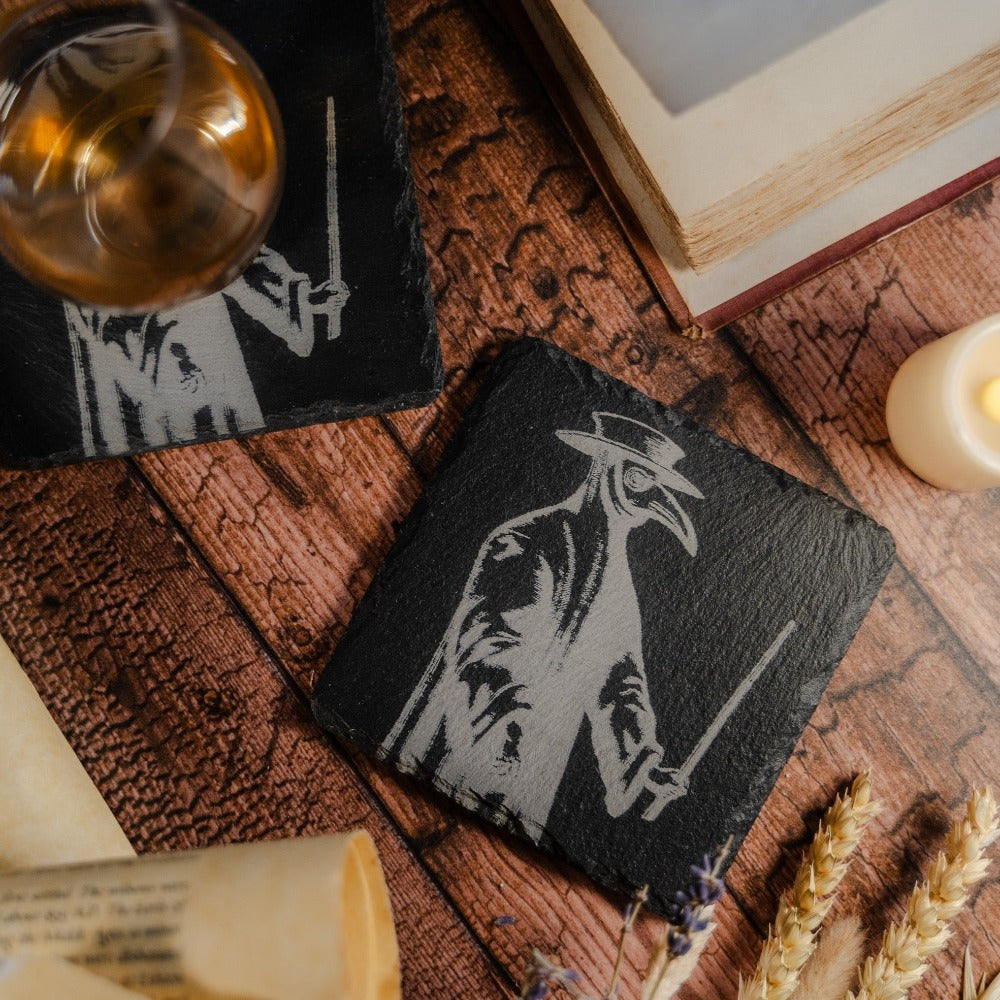 A slate coaster with a charcoal image of a full length plague doctor costume, surrounded by books and whisky