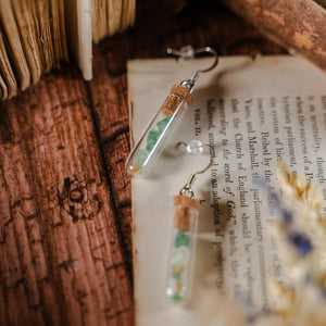 Two test tube earrings filled with green gem stones, laid against dried flowers and an open book