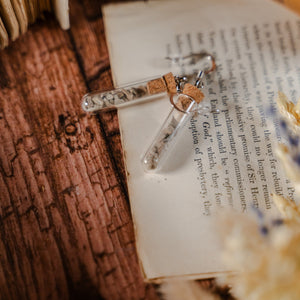Two test tube earrings filled with stone pieces, laid against dried flowers and an open book