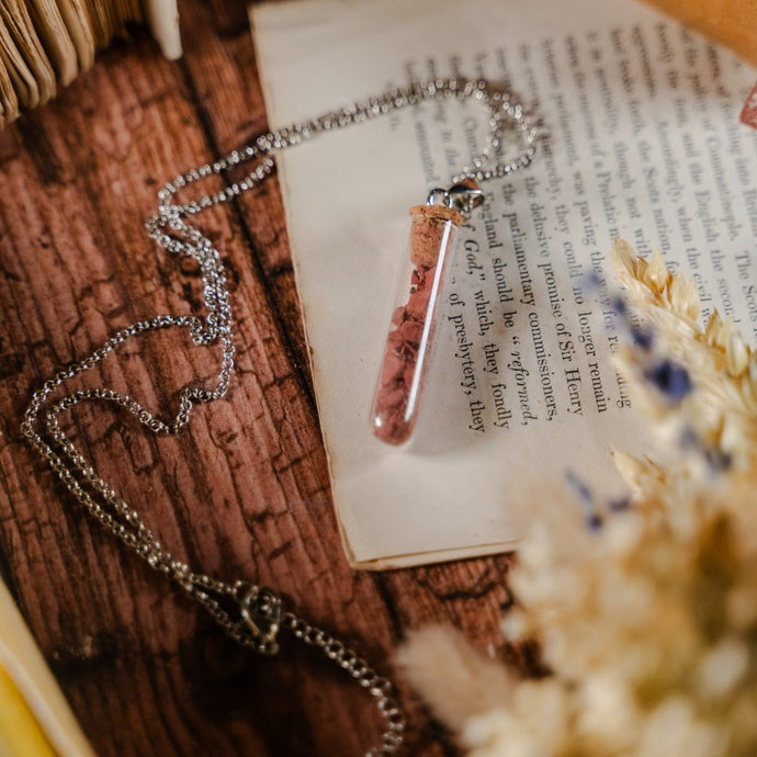 Test tube necklace filled with colourful stones, laid against dried flowers and an open book